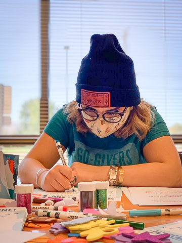 Employee wearing a mask and making handcrafted cards with markers and craft supplies on the table.