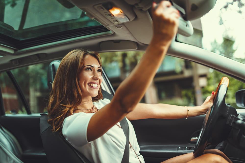 A young happy smiling woman adjusts the car's rear view mirror before driving her new car.